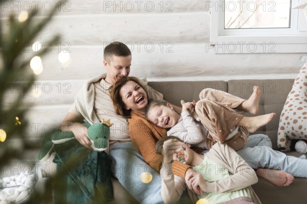 A joyful family celebrates Christmas in a cozy home setting. Two adults and two children, all laughing and cuddling on a sofa, are surrounded by Christmas decorations and soft lighting