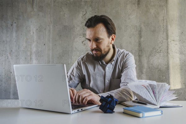 A man in a shirt works intently on a laptop at office. Beside him are a book and a Rubik cube, suggesting multitasking or problem solving in a business setting
