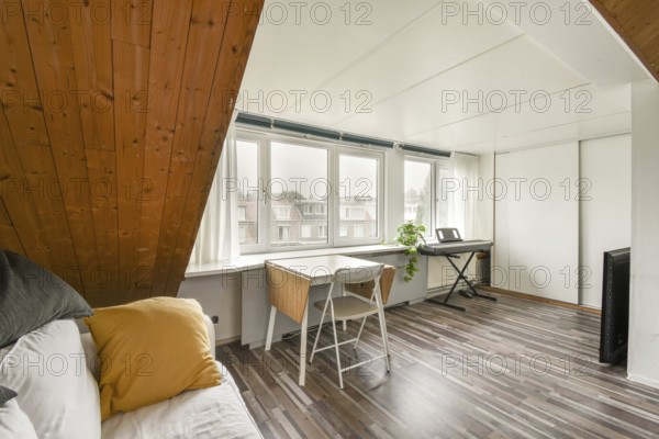 Spacious attic room featuring a slanted wooden ceiling, bright windows with a view, a cozy workspace with a white desk, and a keyboard, creating a perfect creative setting