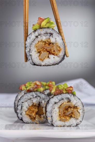 A close up of a sushi roll with vibrant avocado and salmon topping, held by chopsticks with more rolls above a white background. Perfect for food and restaurant themes
