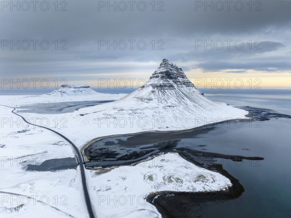 A breathtaking aerial view of Kirkjufell mountain in Iceland, covered in snow, surrounded by icy waters. The tranquil winter landscape contrasts against the dramatic skies