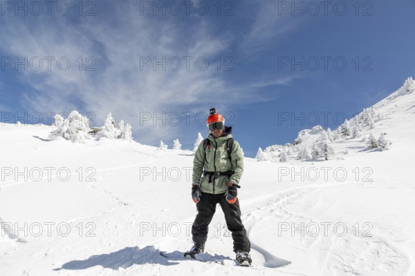A male snowboarder stands confidently in a pristine snowy landscape under a clear blue sky, wearing a green jacket and orange helmet