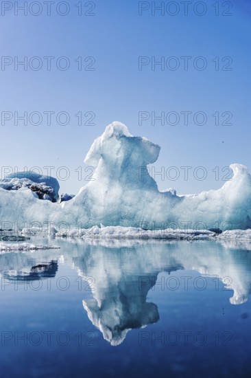 A majestic ice formation towers over the serene waters of Glacier Jokulsarlon in Iceland, reflecting its intricate shapes in the clear, still lagoon under a bright blue sky