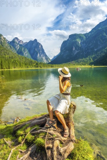 Young woman wearing a straw hat sitting on a tree stump by lake dobbiaco, enjoying the breathtaking view of the dolomites on a summer day