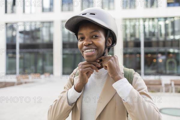 A black woman smiles while adjusting her helmet in an urban setting She is dressed modernly, embodying confidence and style against a cityscape backdrop