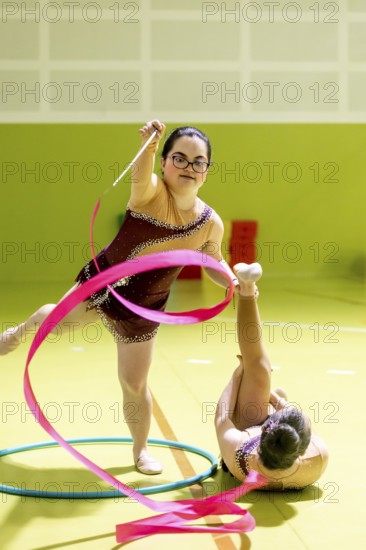 Two disabled rhythmic gymnasts perform with a pink ribbon and hoop in a vibrant indoor space. One stands, the other is on the floor, showcasing athleticism and coordination
