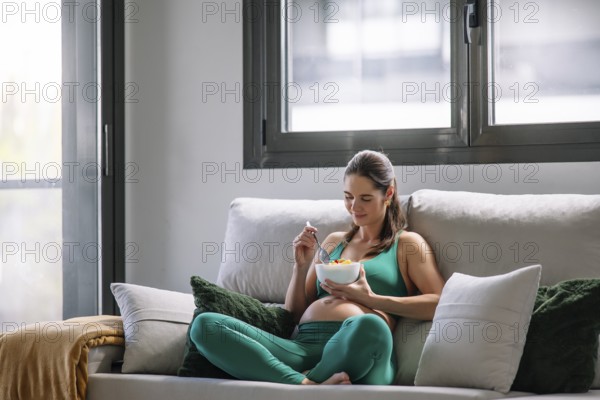 A pregnant woman sits comfortably on a couch in a yoga outfit, enjoying a relaxing moment while eating. Natural light from the window enhances the serene atmosphere of wellness