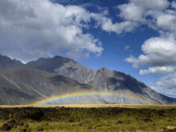 A stunning autumn scene at Mount Cook, New Zealand, showcasing a vibrant rainbow arching over lush green fields against majestic mountains, under a partly cloudy sky