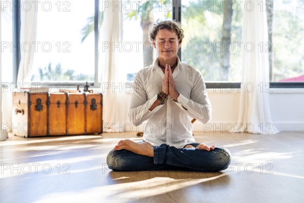 A calm man with curly hair meditates with eyes closed, seated in a lotus pose, hands in prayer position in a sunlit room decorated with minimalistic style and large windows