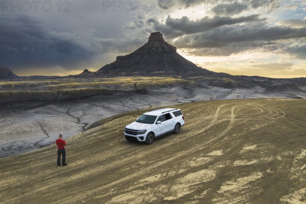 From above back view of unrecognizable man standing near a white SUV at Factory Butte, Utah, as the sun sets over the rugged landscape, showcasing dramatic skies and intricate ground patterns