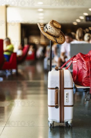 White suitcase adorned with a cowboy hat, indicating travel and adventure, located in a busy airport setting