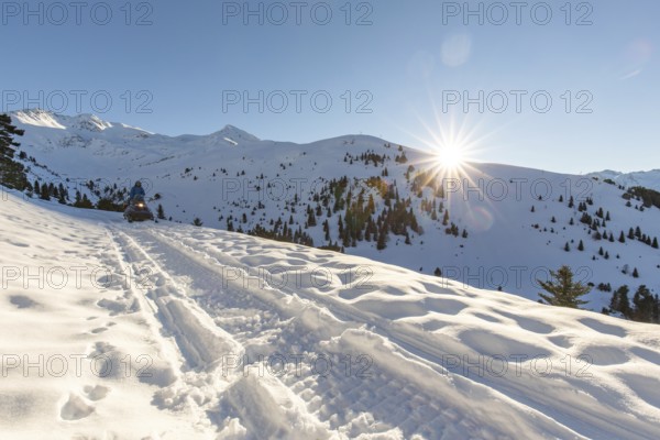 A snowmobile traverses a snowy mountain path under a clear blue sky in a picturesque winter wonderland. The sun shines brightly, casting long shadows across the pristine landscape