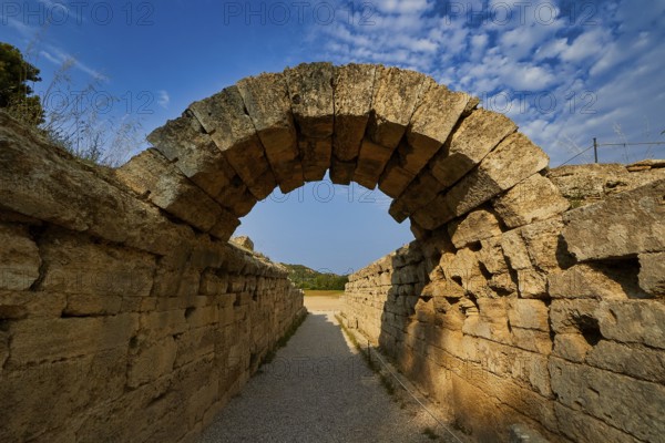 Crypt, vaulted walkway to the stadium, old stone arch leading through massive walls under a blue cloudy sky, Archaeological Site, Ancient Olympia, Peloponnese, Greece