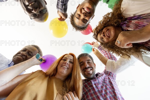 A cheerful group of friends looks down at the camera, holding colorful balloons, symbolizing a lively birthday celebration. Faces are filled with joy and excitement