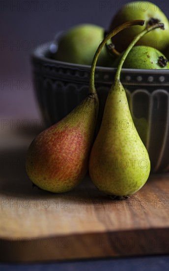 Close up of two wild pears resting on a wooden board with a decorative ceramic bowl filled with more pears in the background, showcasing their natural hues in soft lighting