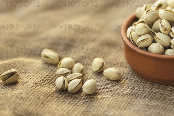 Close up of a clay bowl filled with pistachios on a burlap surface. Several pistachios are scattered around, highlighting the rich texture and warm tones