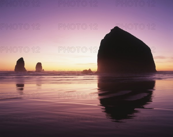 A serene sunset graces the horizon, illuminating ocean waters and silhouetting majestic rock formations. The tranquil scene evokes a sense of peace and natural beauty