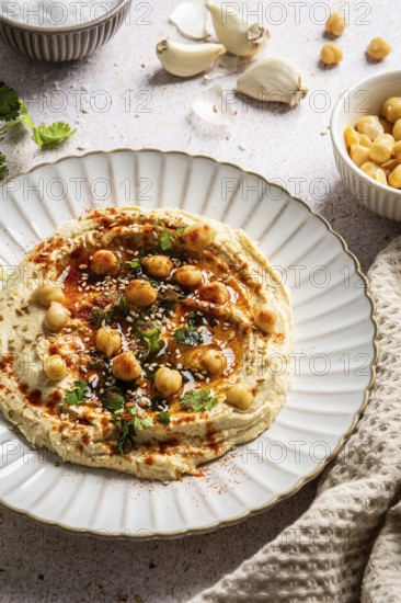 A delicious hummus dish is presented on a decorative plate, topped with chickpeas, sesame seeds, and herbs. Surrounding elements include garlic and a cozy cloth overlay