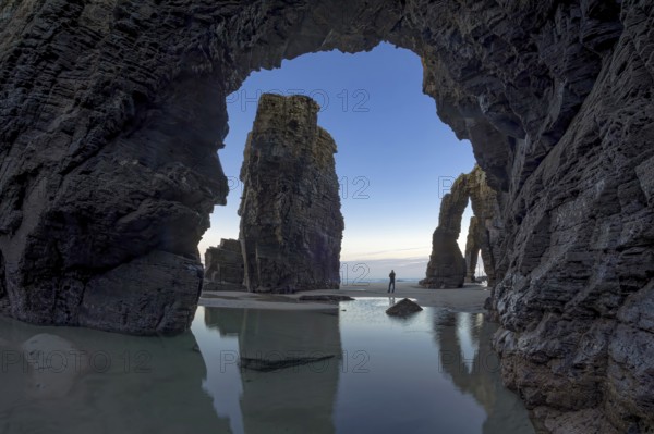 A person stands between towering sea arches, reflected in calm tidal pools, under a serene sky. The scene captures the wonder of nature's architecture