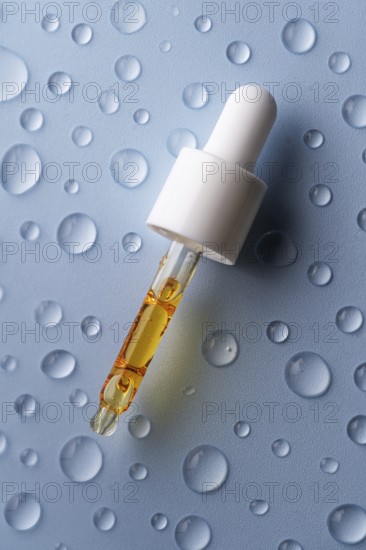 A close-up of a pipette filled with vitamin C serum on a blue surface decorated with water droplets The image highlights a skincare theme with a focus on hydration