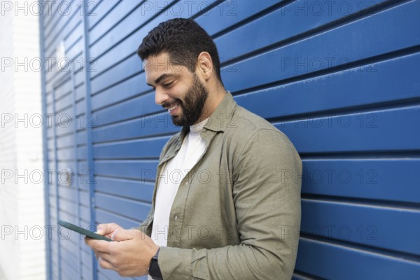 A man stands against a blue wall, smiling as he looks at his smartphone in the city. He wears a casual green shirt and a watch, emphasizing a relaxed and modern lifestyle