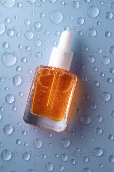 A bottle of vitamin C serum with a pipette, filled with vibrant orange liquid, is set against a blue background with water droplets, highlighting its fresh and hydrating properties