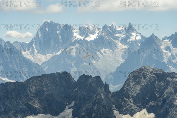 A lone paraglider glides gracefully above the rugged peaks of the French Alps near Chamonix and Mont Blanc, surrounded by dramatic snow-capped mountains under a clear blue sky