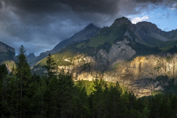 Majestic mountain landscape at Oachinensee, Switzerland, showcasing dramatic clouds and sunlight illuminating the rocky cliffs. Lush green forests enhance the serene alpine scenery
