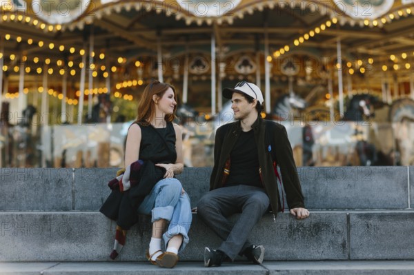 A young couple sits on stone steps, engaged in conversation, with a vibrant carousel in the background in Brooklyn Bridge Park. The warm lights create a cheerful, nostalgic atmosphere