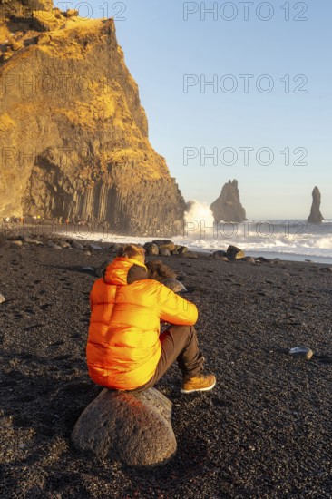 A man in an orange jacket sits on a rock on a beach. The sky is blue and the sun is shining. The man is looking out at the ocean