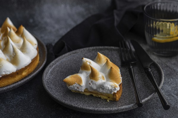 A slice of a homemade lemon cake sits elegantly on a dark plate, accompanied by a fork and knife. A glass with a slice of lemon adds a refreshing touch to the minimalist scene