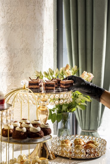 A luxurious dessert spread featuring various cakes and pastries displayed on elegant, crystal adorned stands with a hand reaching for a chocolate treat. Lighting and flowers enhance the ambiance