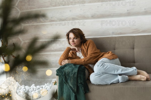 A woman sits cozily on a couch, adorned in a warm orange sweater and light blue jeans, gently looking away while surrounded by Christmas decorations and soft, festive lights