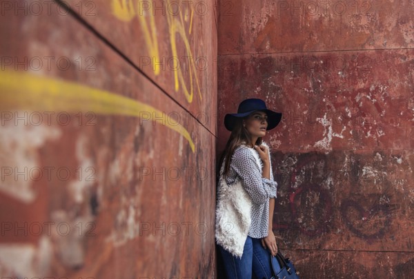 A fashionable woman in a blue hat leans against a weathered red wall adorned with graffiti. She exudes an effortless style, combining modern fashion with an urban backdrop