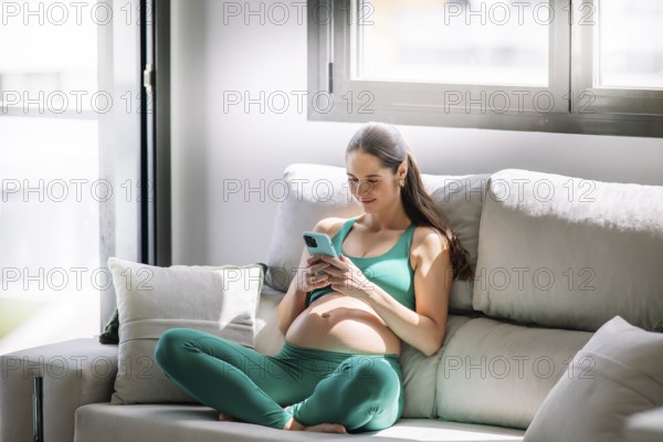 A pregnant woman in a green yoga outfit relaxes on a comfortable couch, focused on her smartphone. The image conveys a sense of calm and wellness during pregnancy