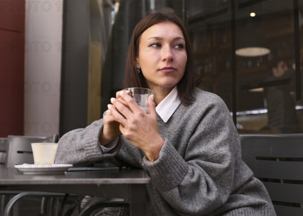 A thoughtful woman in a gray sweater holds a glass while sitting at an outdoor bar table. The cozy setting and reflective mood create a warm, inviting atmosphere