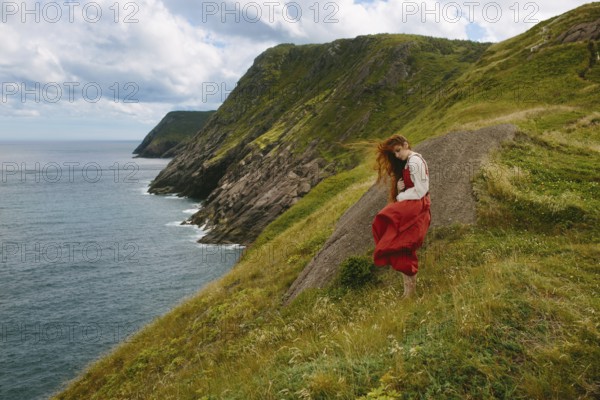 A young woman wearing a flowing red dress and white blouse explores the rugged, green hillside overlooking the ocean at Middle Cove, Newfoundland & Labrador, Canada. Her red hair is tousled by the wind