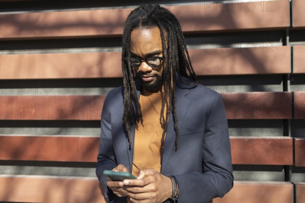 A cuban business professional with glasses and dreadlocks stands outdoors in sunlight, using a smartphone while wearing a smart blazer, against a modern wall backdrop