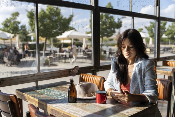 A woman sits alone in a sunlit cafe, looking down at her phone. Natural light flows through large windows, creating a serene atmosphere perfect for relaxation