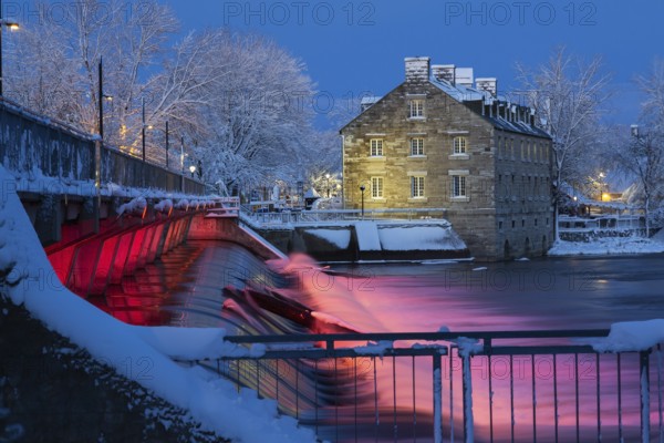 Illuminated Moulin Neuf water flow control dam and walkway over Des Mille-Iles river plus New Mill on Ile des Moulins with first winter like snow in late autumn at dusk, Old Terrebonne, Quebec, Canada