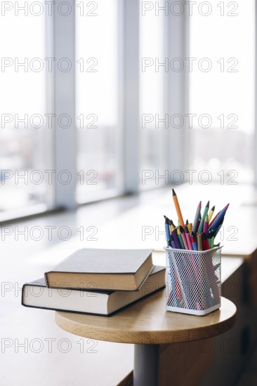 Books with dark covers stacked atop a circular wooden table beside a mesh pencil holder filled with colorful pencils, placed near a large window letting in natural light