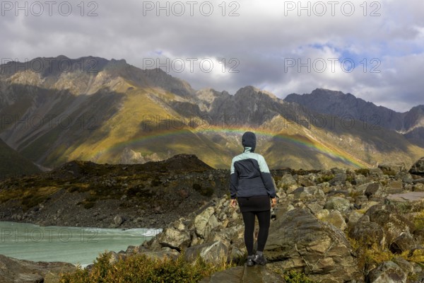 A woman stands on rocky terrain, admiring a vibrant autumn landscape on Mount Cook, New Zealand, with a rainbow arching over majestic mountains under a cloudy sky