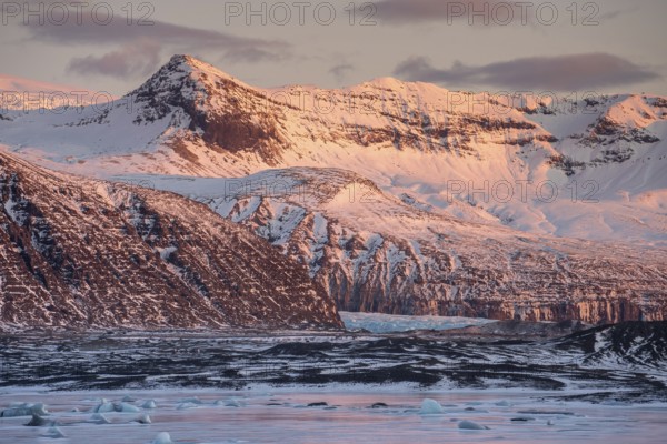 A breathtaking winter sunrise casts a warm glow over the snow-capped mountains and icy landscape in Iceland, capturing the serene beauty and grandeur of the scene in Skaftafell