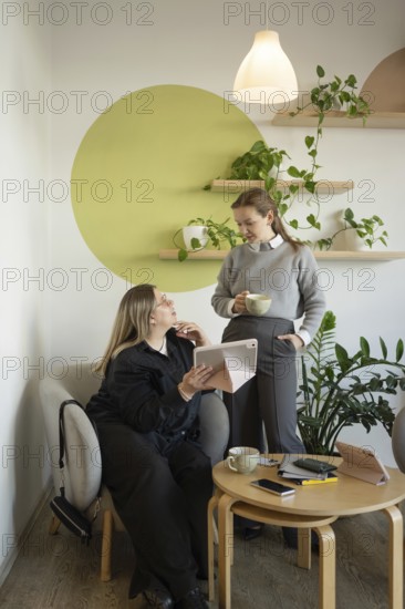 Two women engage in conversation in a cozy cafe setting. One sits with a tablet while the other holds a mug. Aesthetic decor with plants and wall art adds charm