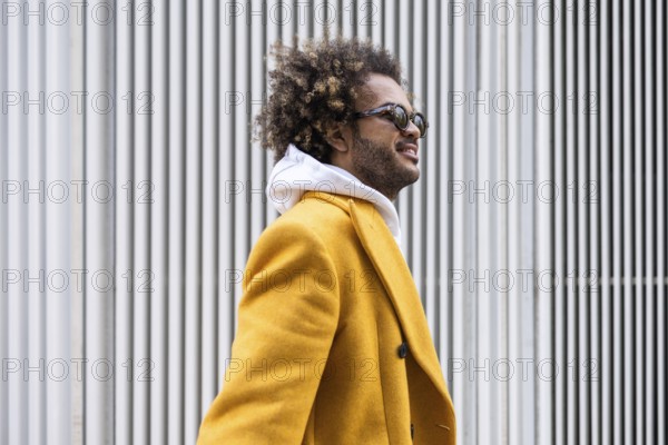 Afro man wearing a stylish mustard coat gazes confidently to the side. His sunglasses reflect a sense of cool sophistication against a backdrop of white vertical lines