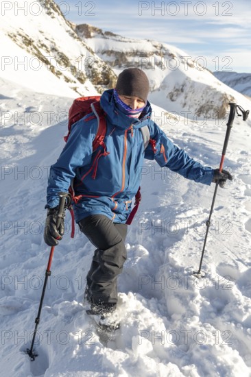 Adventurer in blue jacket and red backpack enjoys ski touring on a sunny day. Using poles for balance, trekking through fresh snow in a mountain landscape