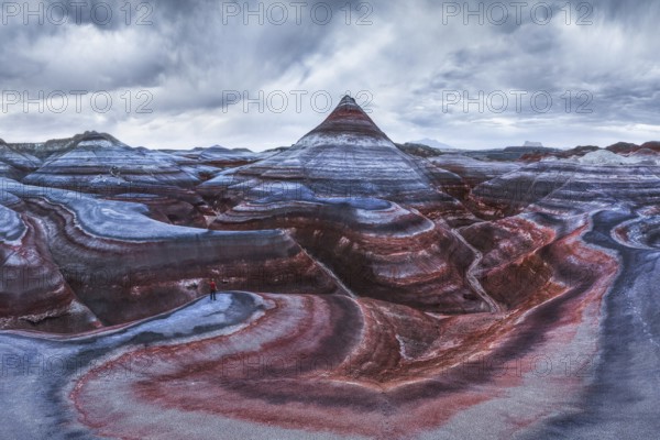 From above aerial drone photograph of an unrecognizable traveler standing on the vibrant and textured geological layers of Hanksville, Utah's landscape under a cloudy sky