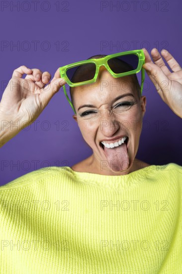 A vibrant middle aged woman with a shaved head wearing bright green sunglasses and a yellow sweater. She playfully sticks out her tongue against a purple backdrop