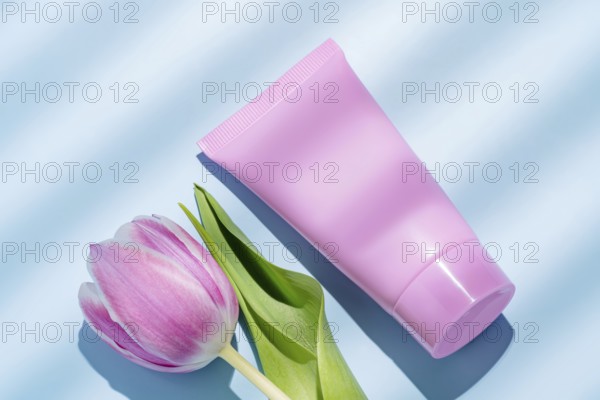 Top view of a vibrant pink cosmetic tube alongside a delicate pink tulip, perfectly arranged on a pale blue background. Ideal for beauty and skincare promotions