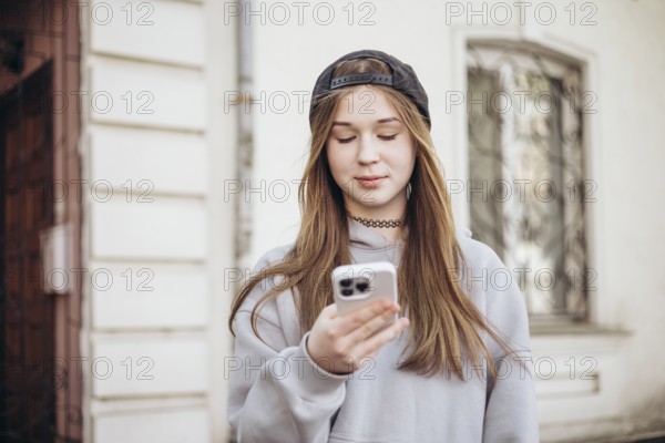 A teenage girl wearing a hoodie and cap is focused on her smartphone while standing outside. She is engaged with her device, representing modern youth culture and technology use
